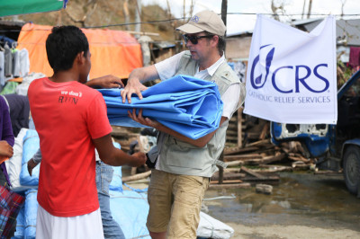 Photo by Jim Stipe/CRS CRS staffer Ross Tomlinson hands out plastic tarps at a CRS and Caritas Norway distribution of 700 emergency shelters in central Philippines. The area was struck by Typhoon Haiyan on November 8, 2013. Jim Stipe/CRS