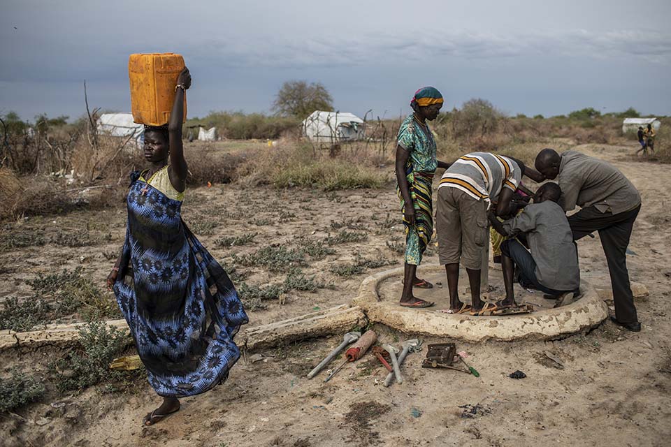 woman carrying jerry can walking past well pump mechanics at work in South Sudan