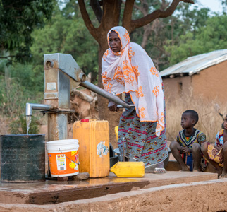 Woman pumping water from on a well with children surraoung her.