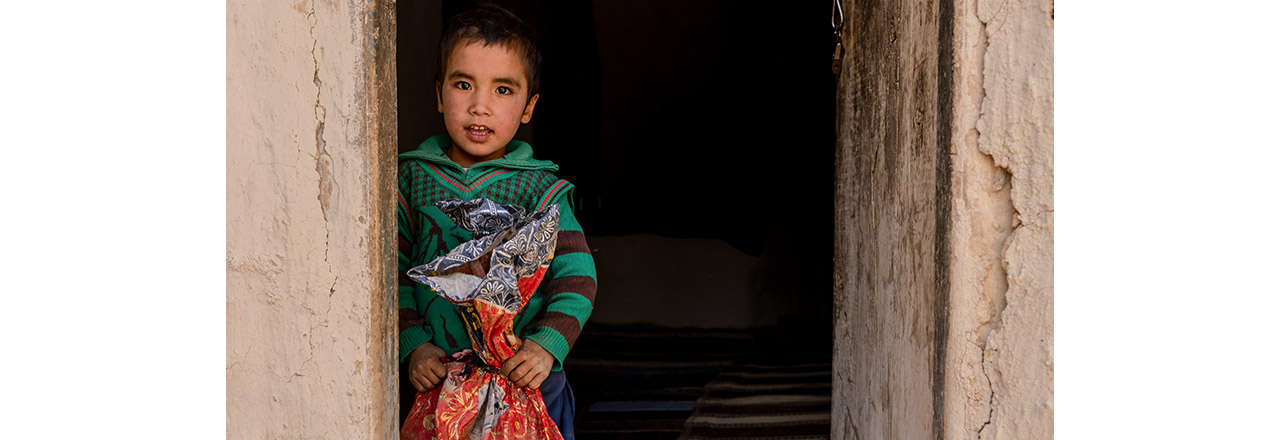 Young Afghani boy stands in doorway holding large bag of rice