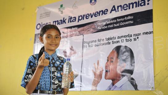 A girl standing in front of a poster about preventing anemia holding a water bottle and a pill.