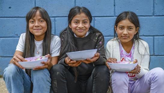 Three smiling girls sitting in front of a blue wall hold empty bowls in their hands after receiving a meal.