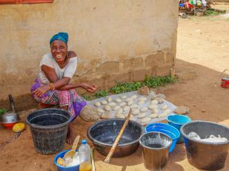 Savings group member sits outside among materials she uses to make shea butter.