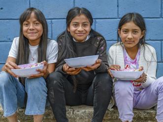 Three smiling girls sitting in front of a blue wall hold empty bowls in their hands after receiving a meal.
