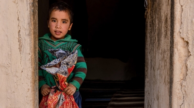 Young Afghani boy stands in doorway holding large bag of rice