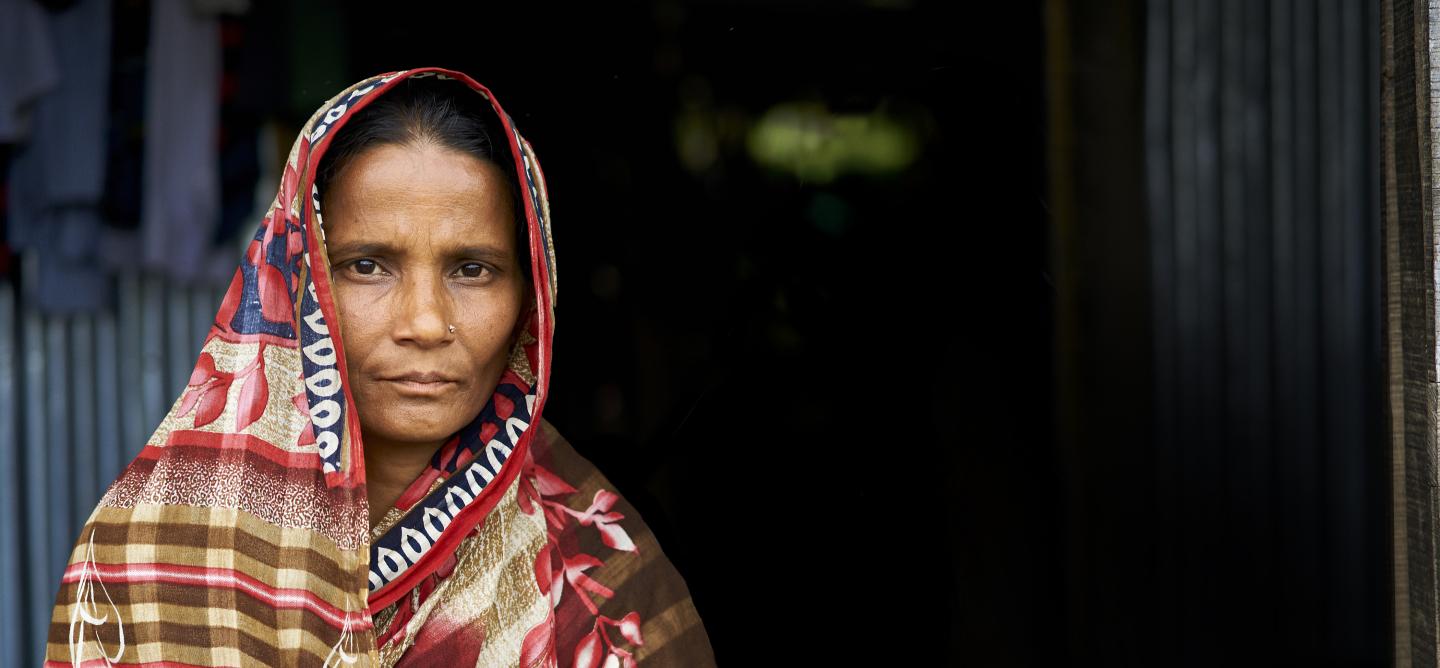 A woman in a patterned saree stands in front of a dark doorway, looking directly at the camera with a neutral expression.