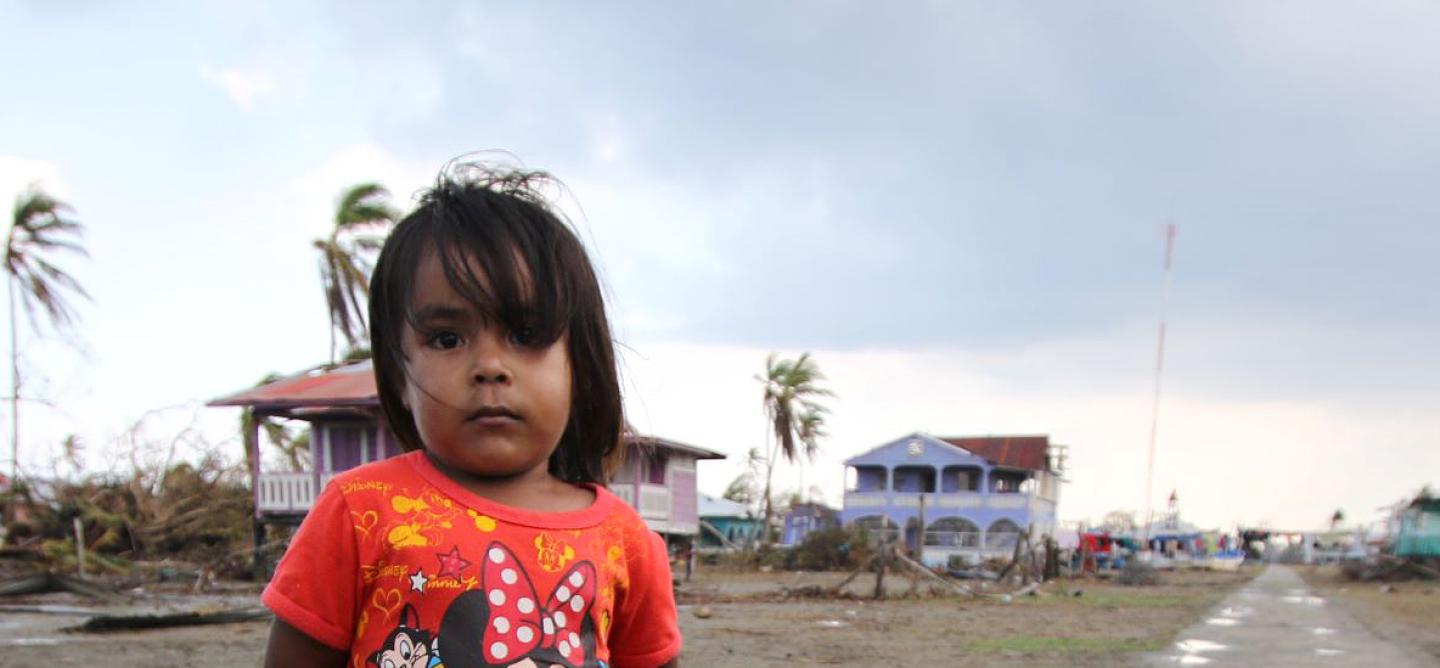 Young girl in a red Minnie Mouse shirt standing on a muddy road in a coastal village damaged by Hurricane Eta, with debris and damaged houses in the background.