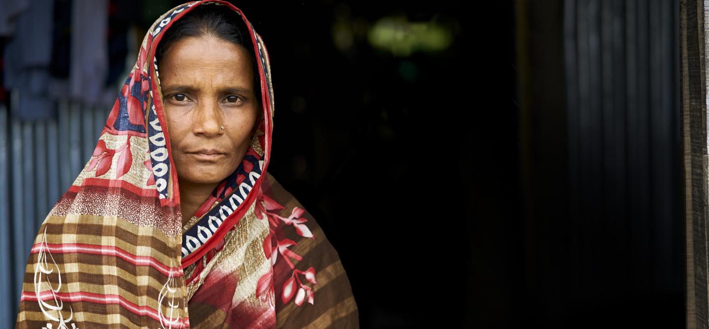 A woman in a patterned saree stands in front of a dark doorway, looking directly at the camera with a neutral expression.