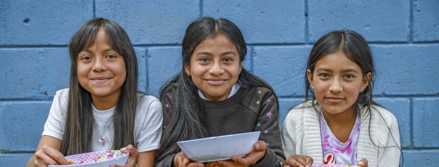 Three smiling girls sitting in front of a blue wall hold empty bowls in their hands after receiving a meal.