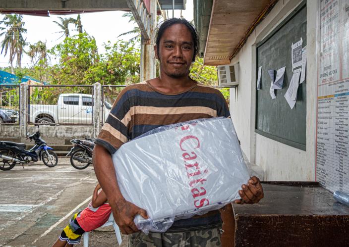 Man holding a plastic-wrapped relief package labeled "Caritas" while standing outside a community building, with motorcycles and trees in the background.