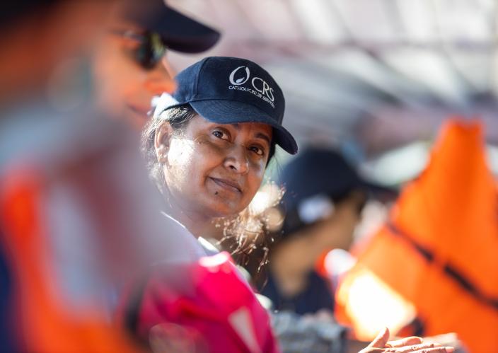 Woman wearing a CRS (Catholic Relief Services) cap looking at the camera, surrounded by others in safety gear, suggesting a humanitarian or relief effort.