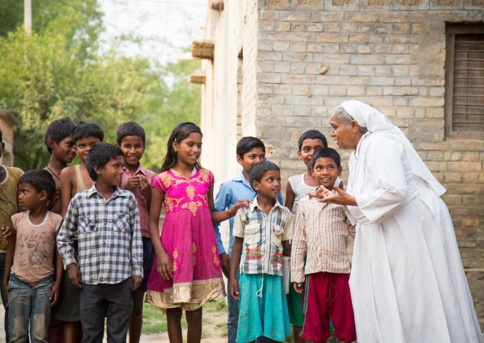 A nun in a white habit warmly interacts with a group of smiling children standing outside a brick building in a rural setting.