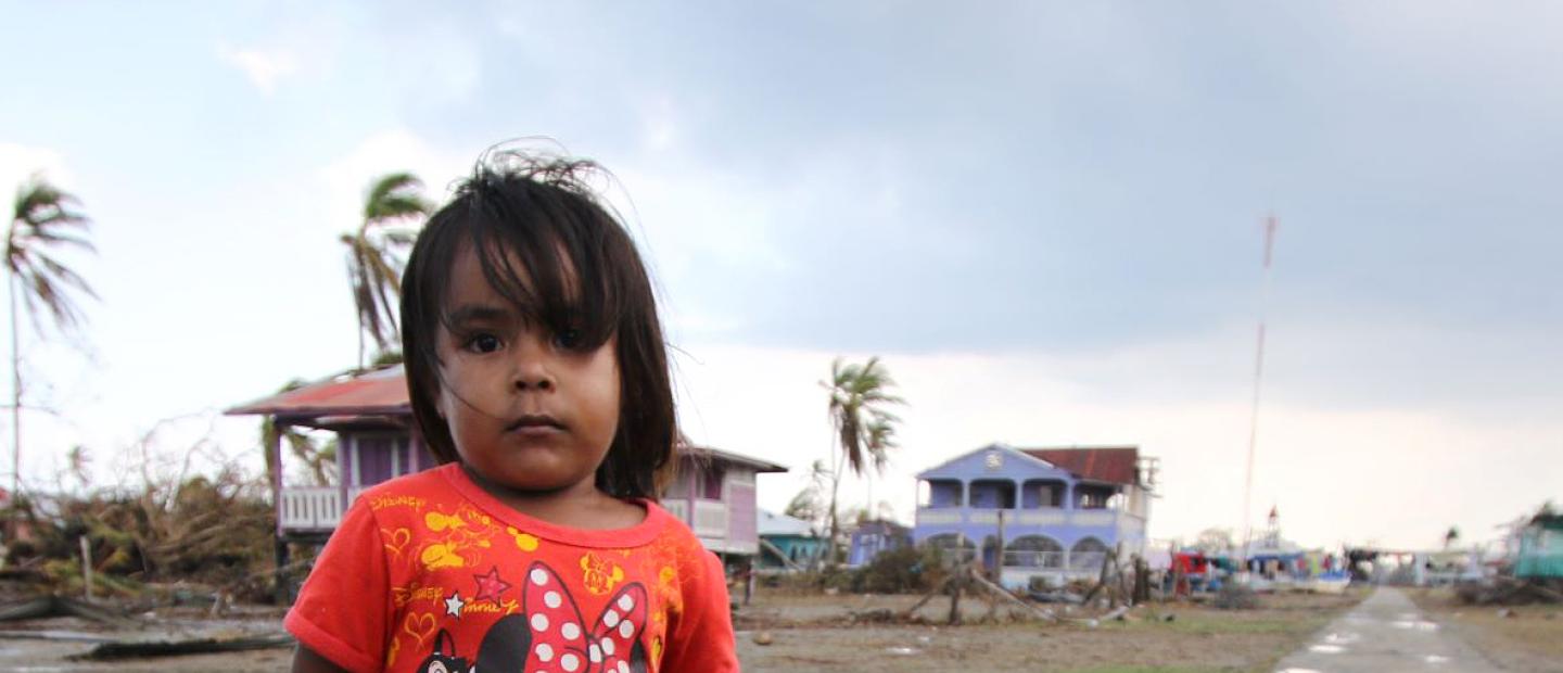 Young girl in a red Minnie Mouse shirt standing on a muddy road in a coastal village damaged by Hurricane Eta, with debris and damaged houses in the background.