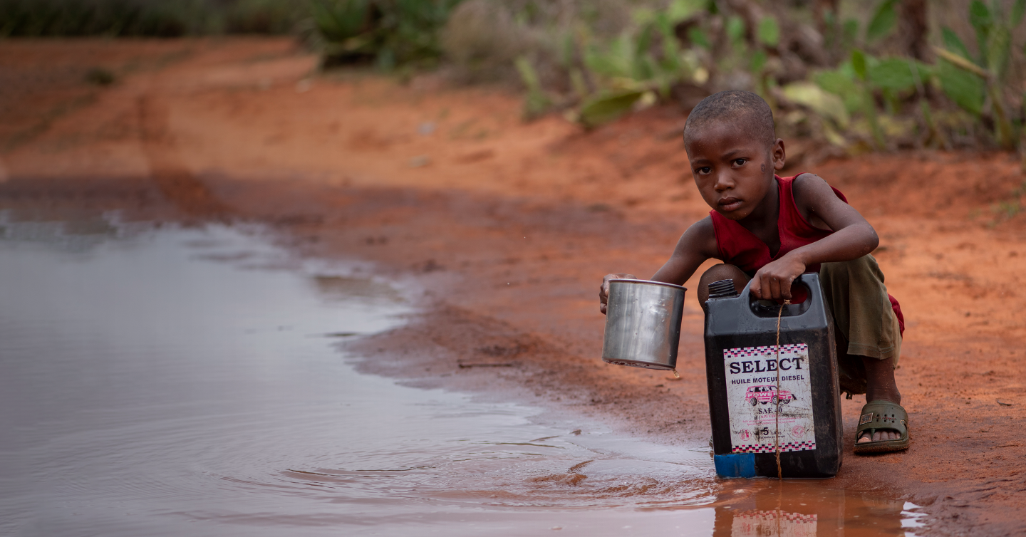 A child crouches by a muddy puddle on a dirt road, using a metal cup to collect water into a plastic container.