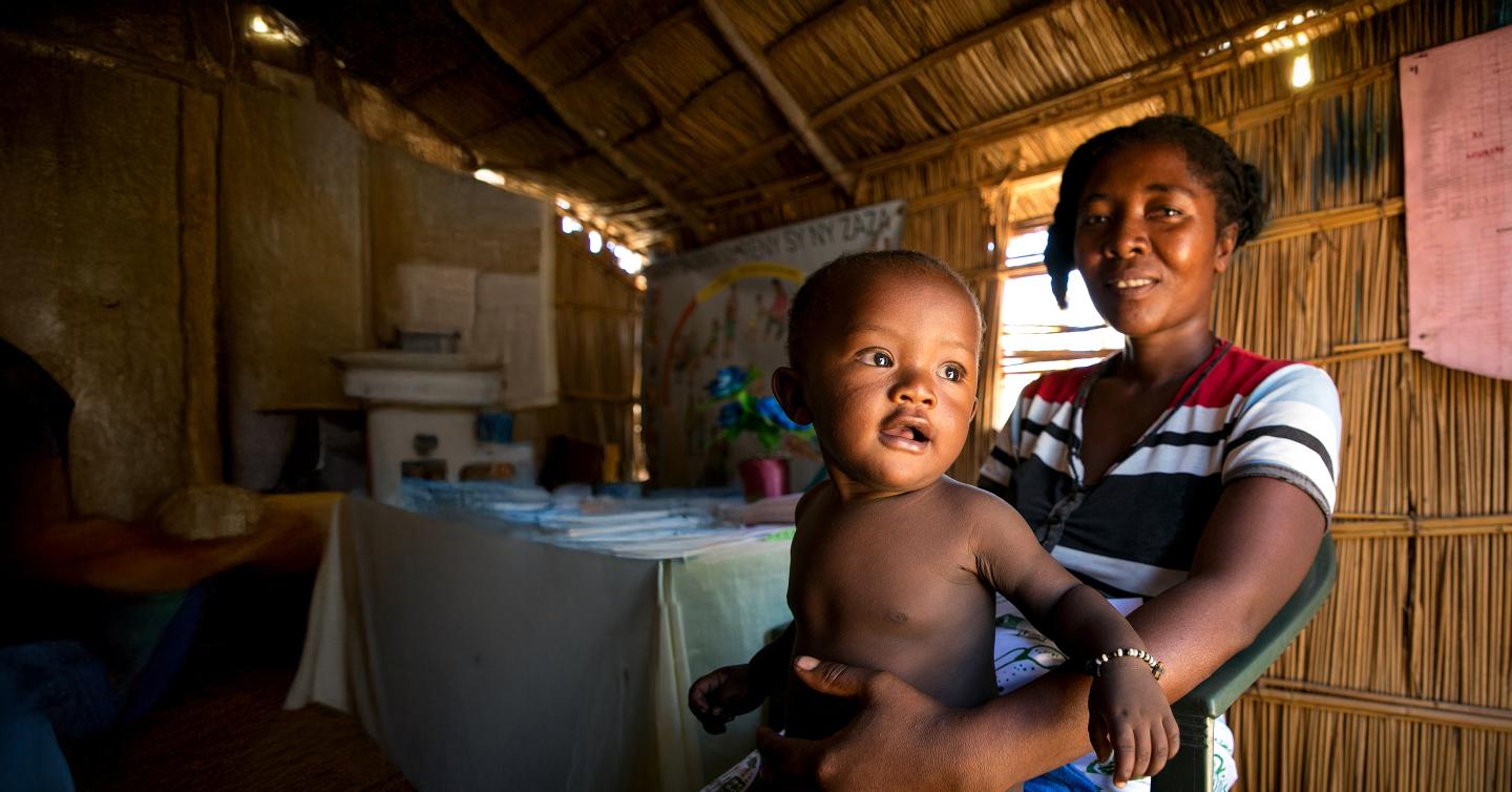 A woman sits inside a thatched-roof home holding her child