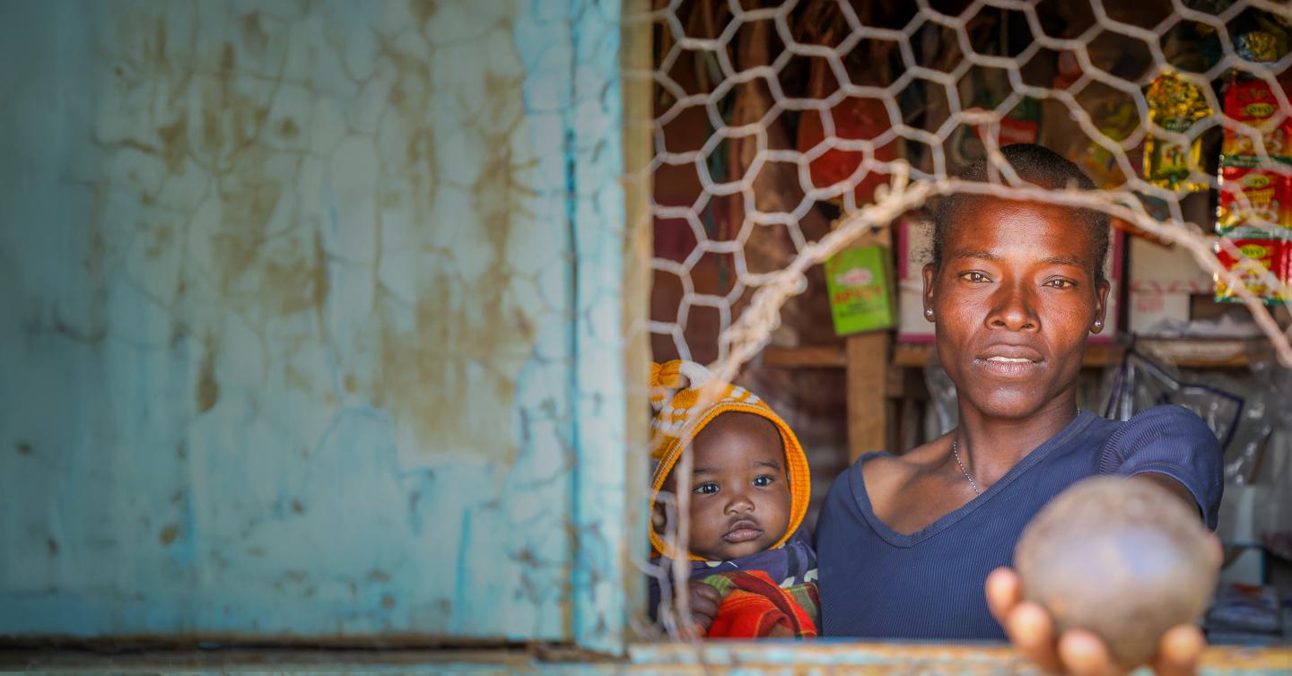 A woman holding a round object through a broken window. On her other arm she's holding a baby.