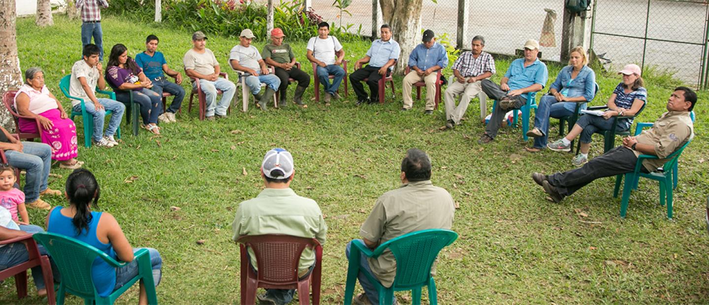 Community members seated in a large circle outdoors, engaged in a group discussion on a grassy area.