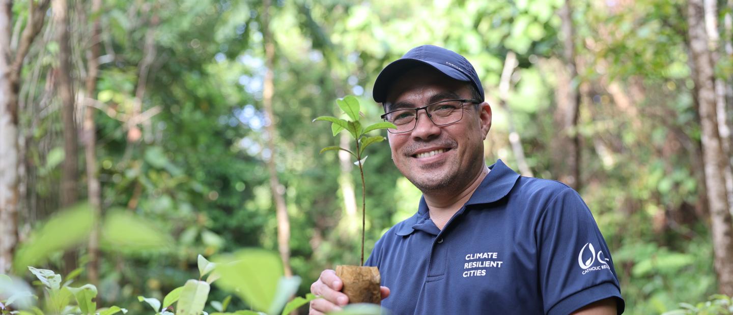 Man in a forest nursery smiling while holding a young tree seedling, wearing a “Climate Resilient Cities” shirt and cap.