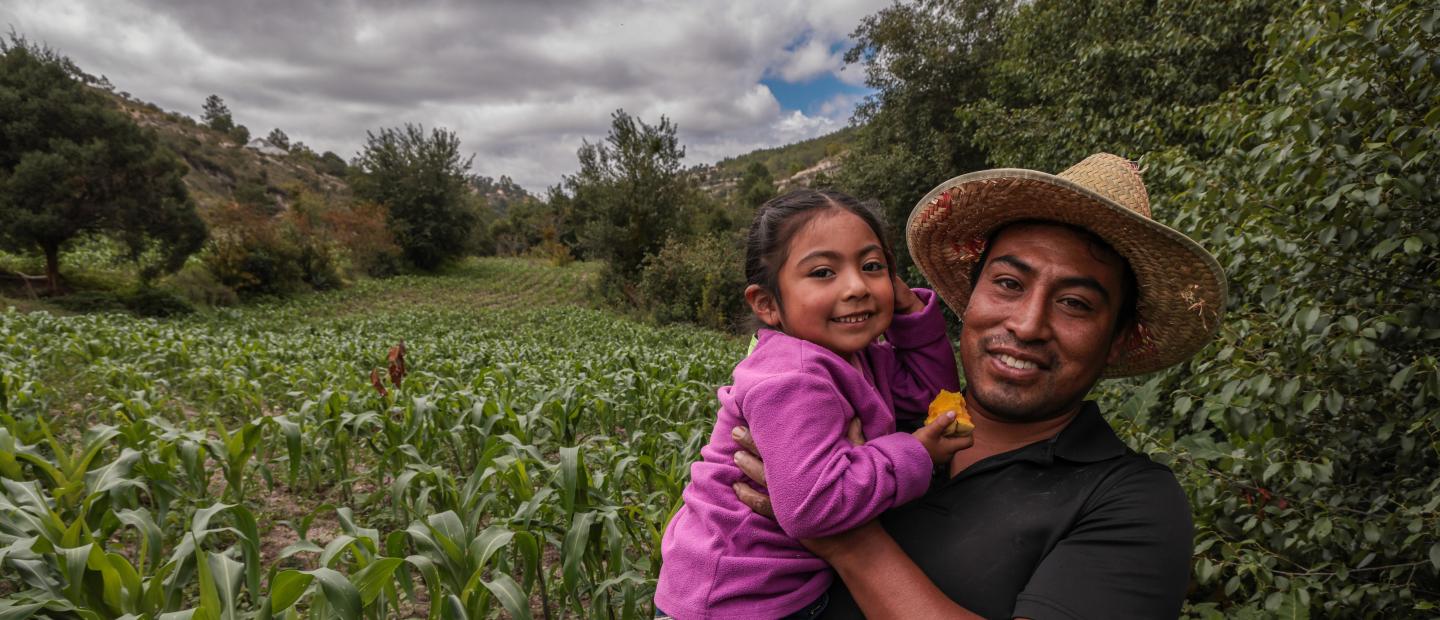 A man holding his daughter in front of their corn field.