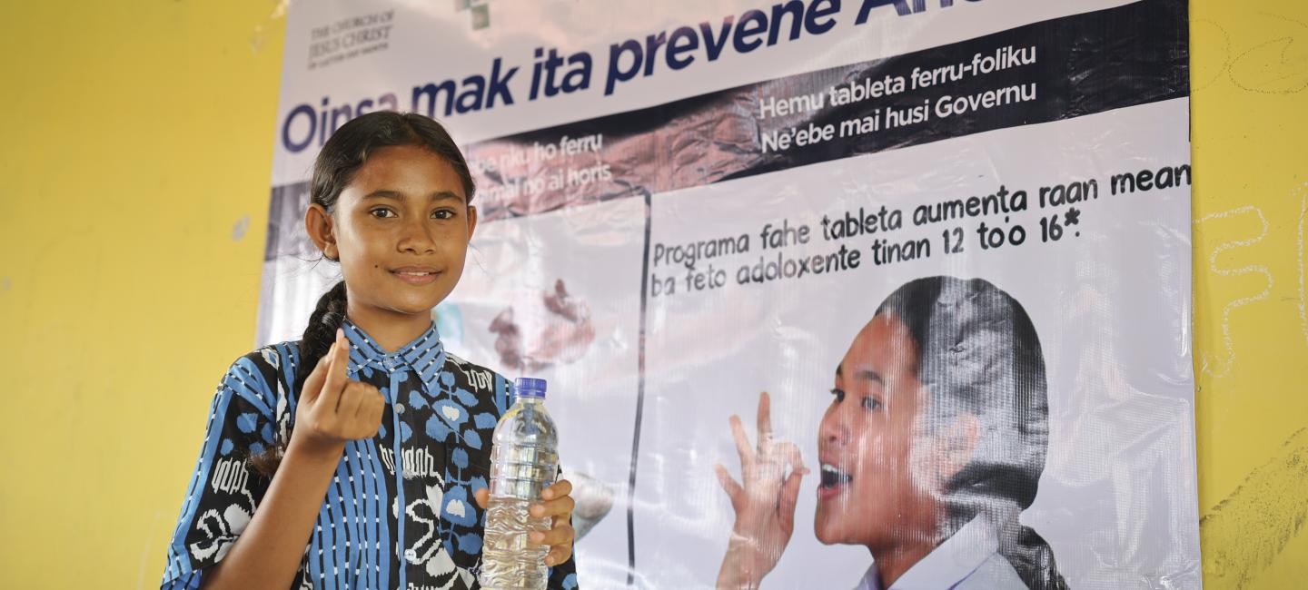 A girl standing in front of a poster about preventing anemia holding a water bottle and a pill.