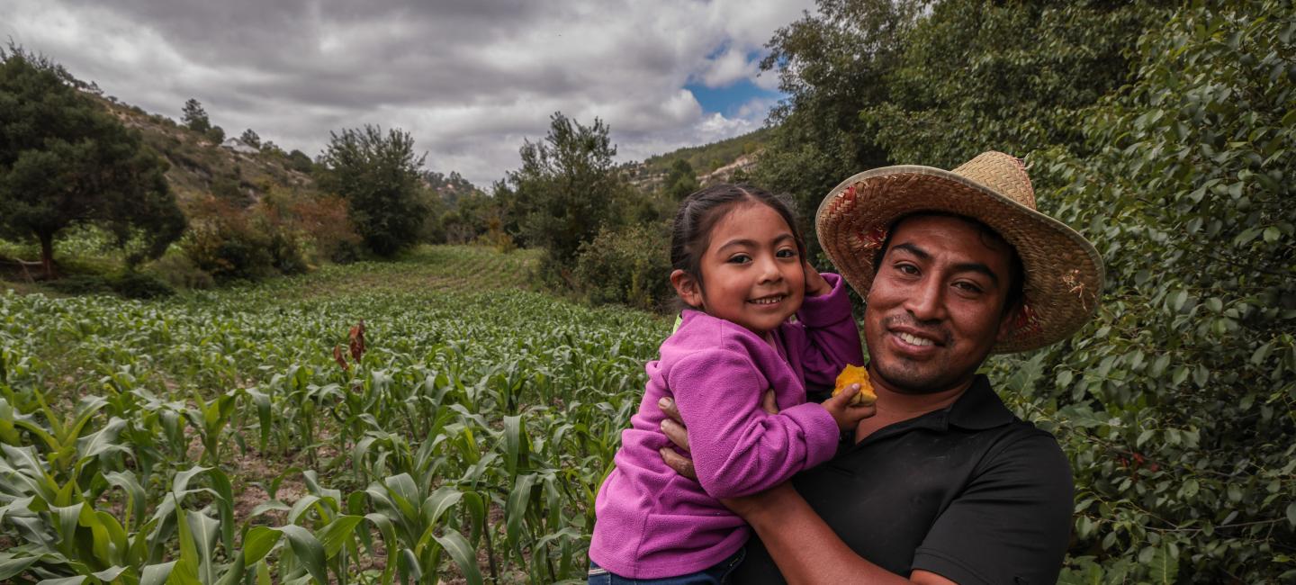A man holding his daughter in front of their corn field.