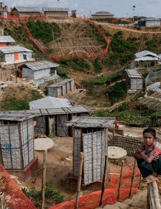 young girl sits on a fence by homes on a hillside at a refugee camp