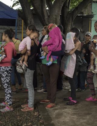 Refugees stand in a line holding their belongings and infant children
