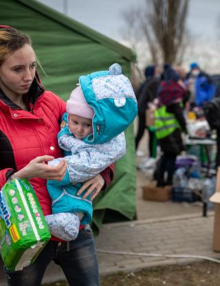Young mother in red coat holds a pack of diapers and an infant in a blue coat.