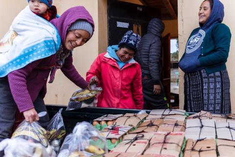 2 farmers with child unload their produce for sale as customer watches.