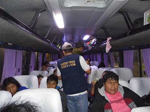 Passengers inside a dimly lit bus, with an official wearing a vest labeled "Ministerio Público Fiscalía de la Nación" speaking to them.