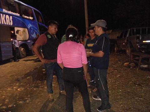 A group of people gathers at night near a parked bus on a dirt road, engaged in conversation with an official wearing a cap and dark clothing.