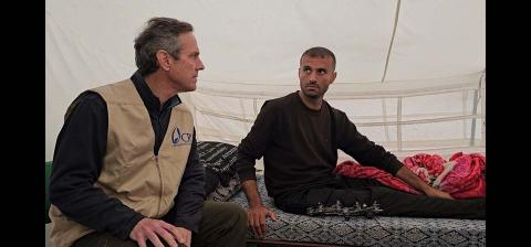 A man sitting by an injured man inside a tent.
