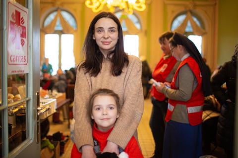 Mother in tan sweater stands behind young daughter in red sweater.