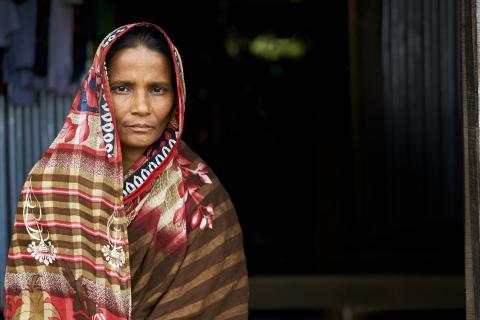 A woman in a patterned saree stands in front of a dark doorway, looking directly at the camera with a neutral expression.