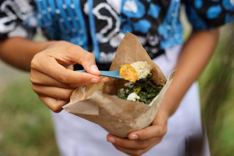 Hands holding a brown paper container of food, using a blue plastic spoon to scoop rice and vegetables.