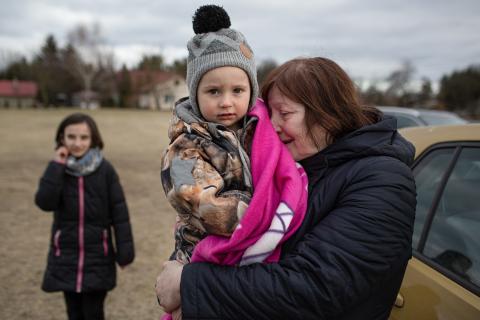 An older woman, appearing emotional, hugs a young boy wrapped in a pink blanket outdoors, while a girl stands in the background on a cloudy day.