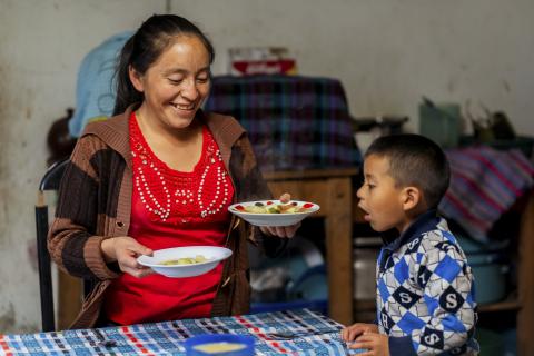 A woman smiling while serving soup to a young boy at a table, both inside a modest, colorful home kitchen.