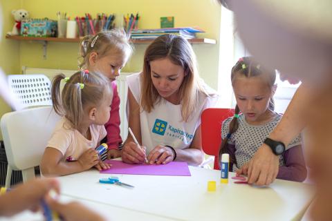 A woman assisting young girls with a craft activity at a table, surrounded by art supplies like scissors, glue sticks, and colored paper in a classroom setting.