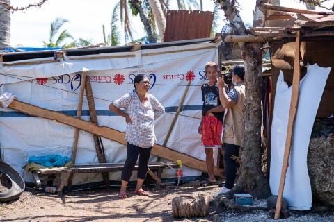 Three people converse outside a makeshift shelter made of tarpaulin and wood, bearing the Caritas relief organization logo. The setting suggests a disaster-affected or impoverished area with improvised housing.