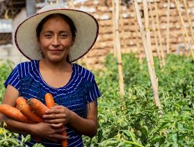 Woman poses in front of farm plot holding bunch of carrots