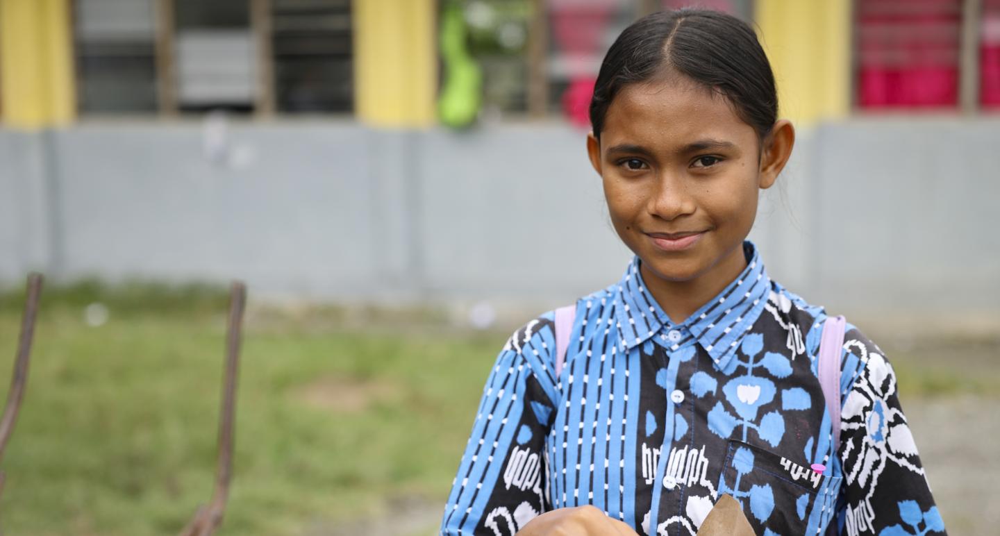 A girl in a blue patterned shirt stands outdoors holding a small brown paper bag