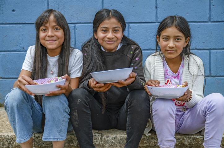 Three smiling girls sitting in front of a blue wall hold empty bowls in their hands after receiving a meal.