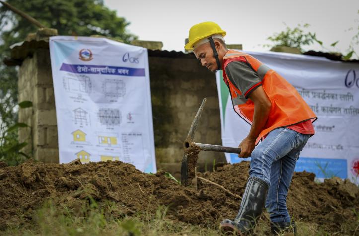 "A man wearing a yellow helmet and an orange vest working on the ground with an excavation tool.