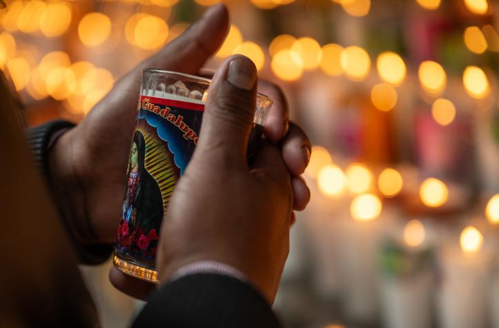 Hands holding a candle with the image of Our Lady of Guadalupe, surrounded by the lights of other lit candles in the background.