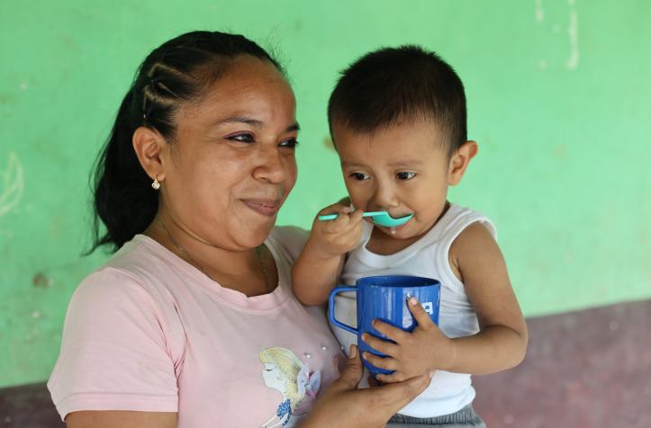 Una mamá carga en brazos a su hijo pequeño mientras él come atol.