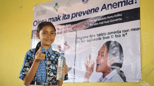 A girl standing in front of a poster about preventing anemia holding a water bottle and a pill.